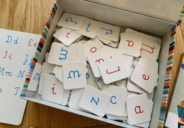 Pudełko z kartami do nauki polskiego alfabetu pisanego. Litery w kolorze niebieskim i czerwonym. Obok biała tablica z zapisanymi literami.