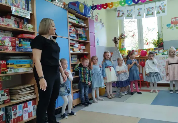 Nauczycielka i dzieci w przedszkolu, na półkach gry i zabawki.