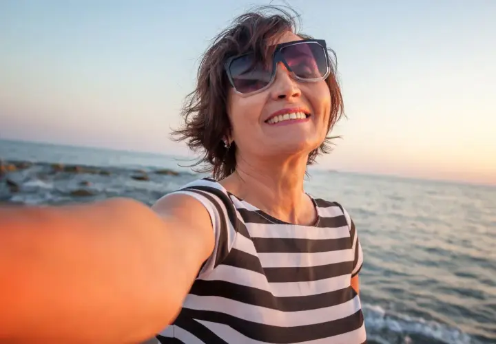 Uśmiechnięta kobieta w okularach przeciwsłonecznych robi selfie na plaży o zachodzie słońca.