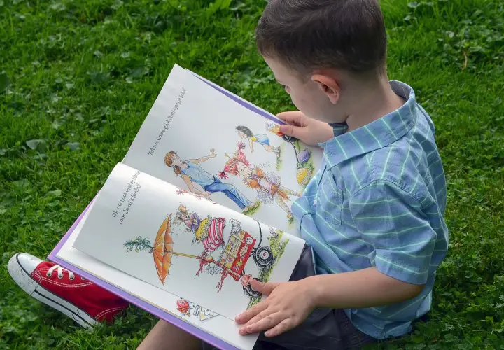 Chłopiec w niebieskiej koszuli czyta otwartą książkę z kolorowymi ilustracjami na trawie.
