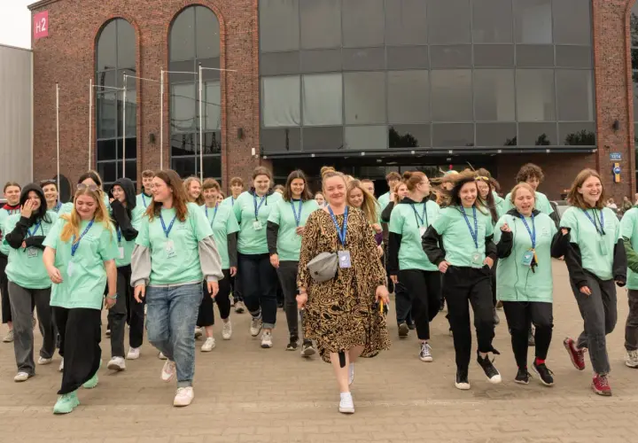Grupa młodych ludzi w zielonych koszulkach idzie przed budynkiem z cegły.