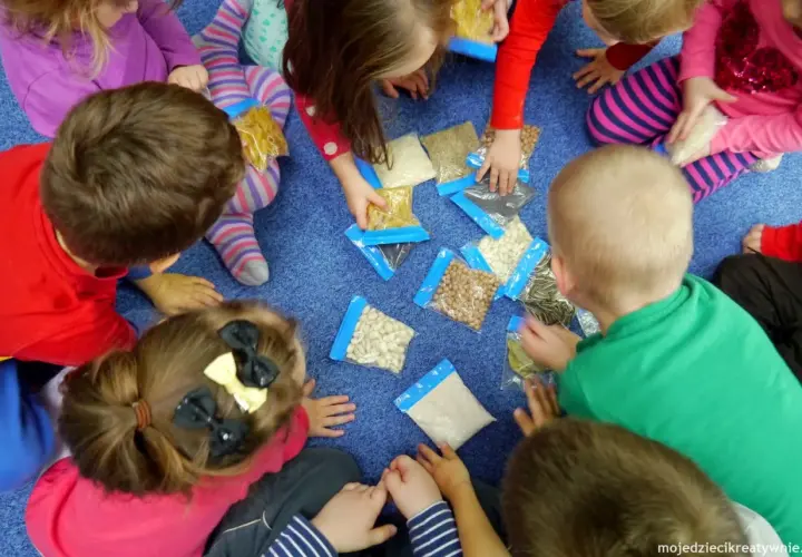 Dzieci siedzą w kółku na niebieskim dywanie, bawiąc się różnymi produktami spożywczymi w woreczkach.