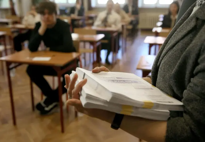 Ręka trzyma stos arkuszy egzaminacyjnych z napisem "JĘZYK POLSKI". W tle uczniowie siedzą przy ławkach.