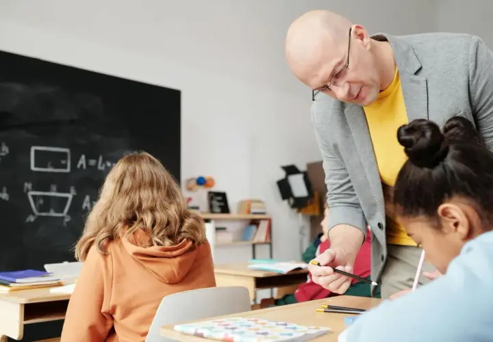 Nauczyciel pomaga uczniom przy biurkach w klasie, na tablicy widać wzory matematyczne.