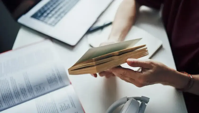 Osoba przegląda książkę, przygotowując się do napisania bibliografii. Obok leży laptop i słuchawki.
