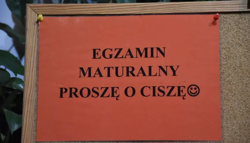 Pomarańczowa kartka na tablicy korkowej z napisem "EGZAMIN MATURALNY PROSZĘ O CISZĘ" i uśmiechniętą buźką.