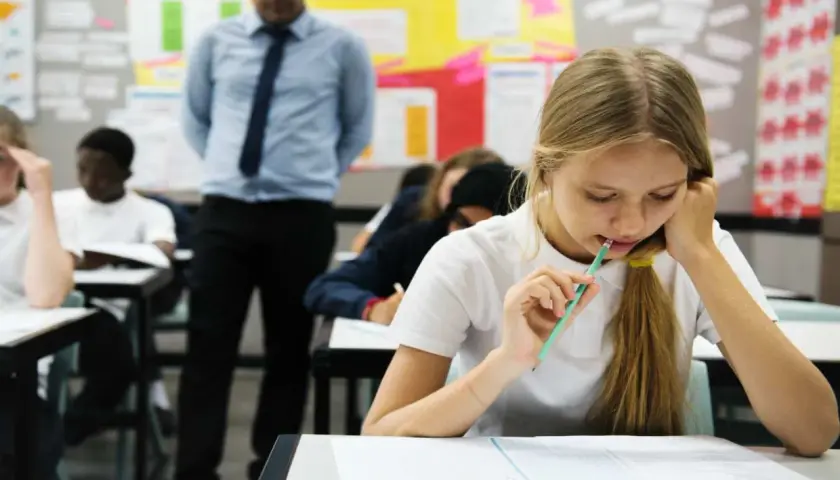 Dziewczynka z ołówkiem w ustach pisze test, nauczyciel stoi z tyłu.