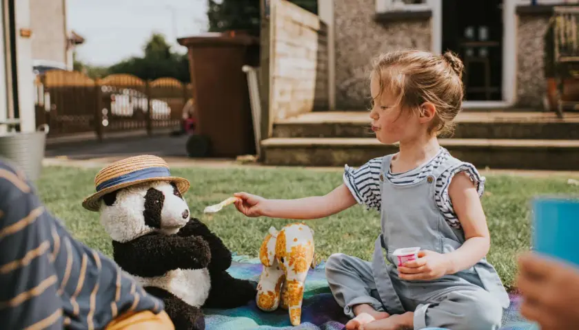 Dziewczynka karmi pluszową pandę w kapeluszu na pikniku na trawie.