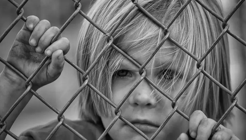Czarno-białe zdjęcie dziecka z blond włosami, patrzącego przez siatkę ogrodzeniową.