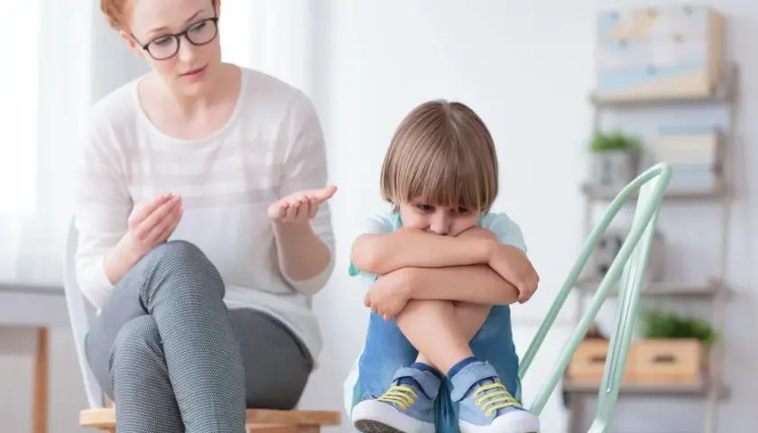Kobieta w okularach rozmawia ze smutnym chłopcem siedzącym na krześle.