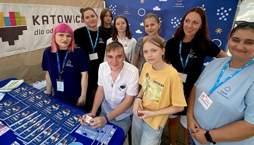 Grupa osób, w tym młodzież i dorosłych, stoi za stołem z książkami i materiałami promocyjnymi "Szkoła w Chmurze".