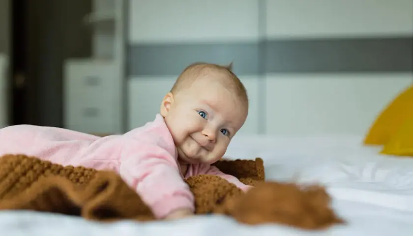 Uśmiechnięte niemowlę w różowym pajacyku leży na łóżku obok brązowego kocyka.