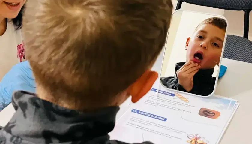 Chłopiec ćwiczy mowę przed lustrem, wspierany przez nauczycielkę.