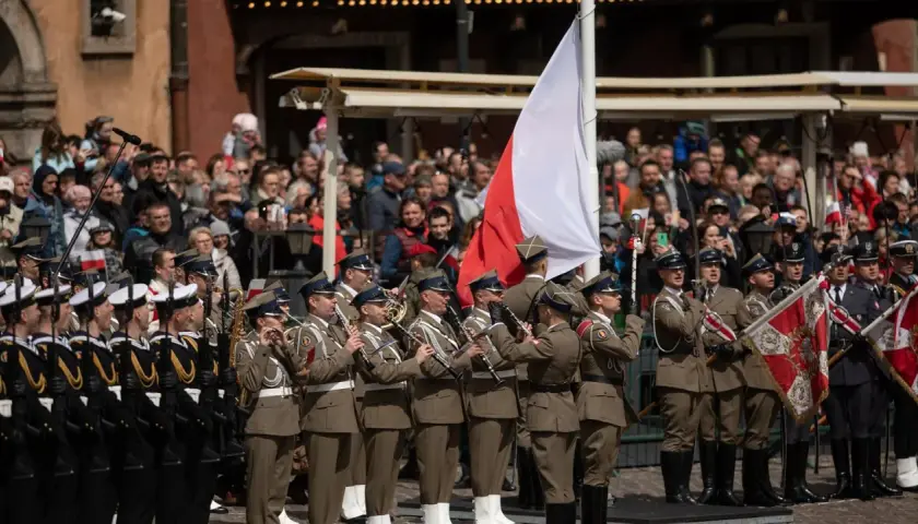 Orkiestra wojskowa grająca na instrumentach dętych na tle tłumu i polskiej flagi.