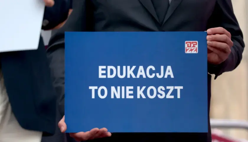 Niebieska tablica z białym napisem "EDUKACJA TO NIE KOSZT" i logo ZZ.