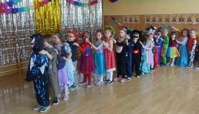 Dzieci w strojach karnawałowych stoją w kolejce na tle błyszczących zasłon i dekoracji.