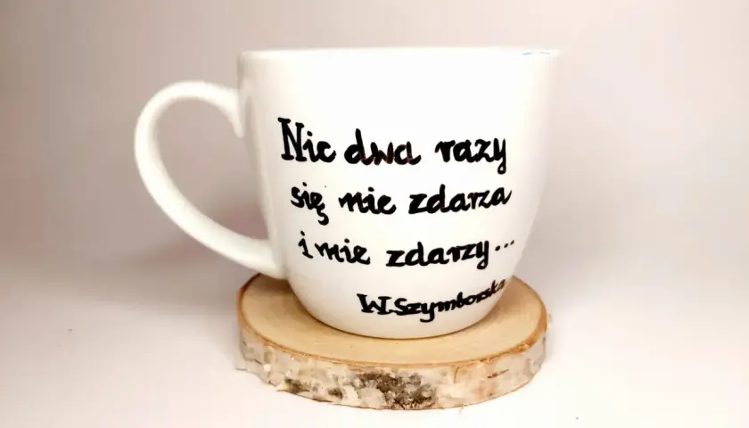 Biały kubek z cytatem Wisławy Szymborskiej "Nic dwa razy się nie zdarza..." na drewnianym podkładzie.