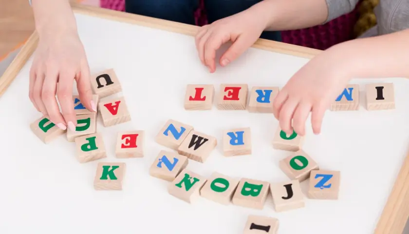 Dzieci układają litery na tablicy, ćwicząc podział na głoski. Widoczne litery to m.in. U, A, T, R, E, N, K, P, Z, O, B, J, I.