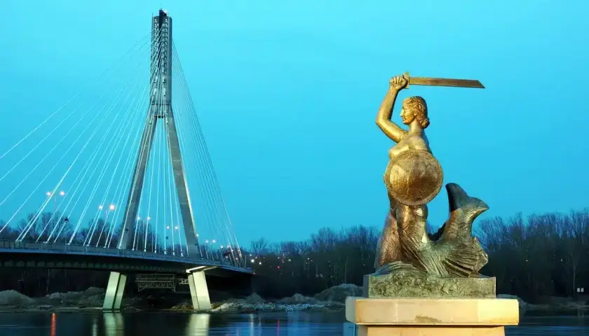 Pomnik Syrenki Warszawskiej z mieczem i tarczą na tle mostu i rzeki.