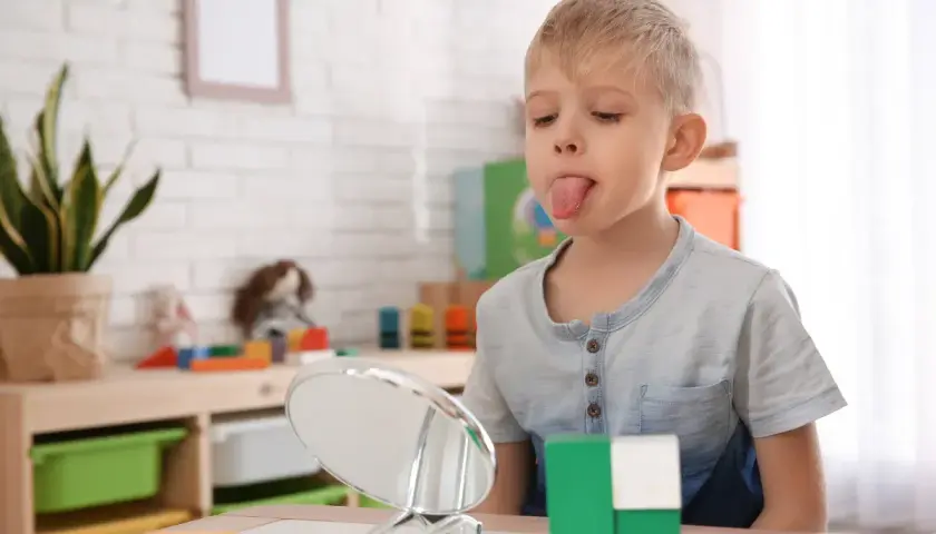 Chłopiec z wysuniętym językiem patrzy w lustro, obok kolorowe klocki.