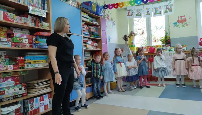 Nauczycielka i dzieci w przedszkolu, na półkach gry i zabawki.
