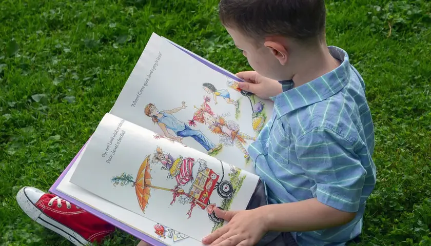 Chłopiec w niebieskiej koszuli czyta otwartą książkę z kolorowymi ilustracjami na trawie.