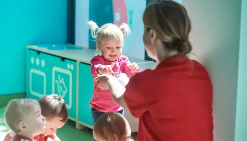 Nauczycielka w czerwonej bluzce bawi się z dziećmi w różowych i czerwonych ubrankach.