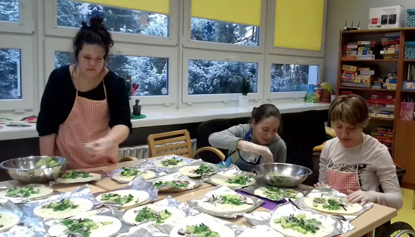 Trzy kobiety w fartuchach przygotowują jedzenie na stole, na tle okien ze śniegiem.