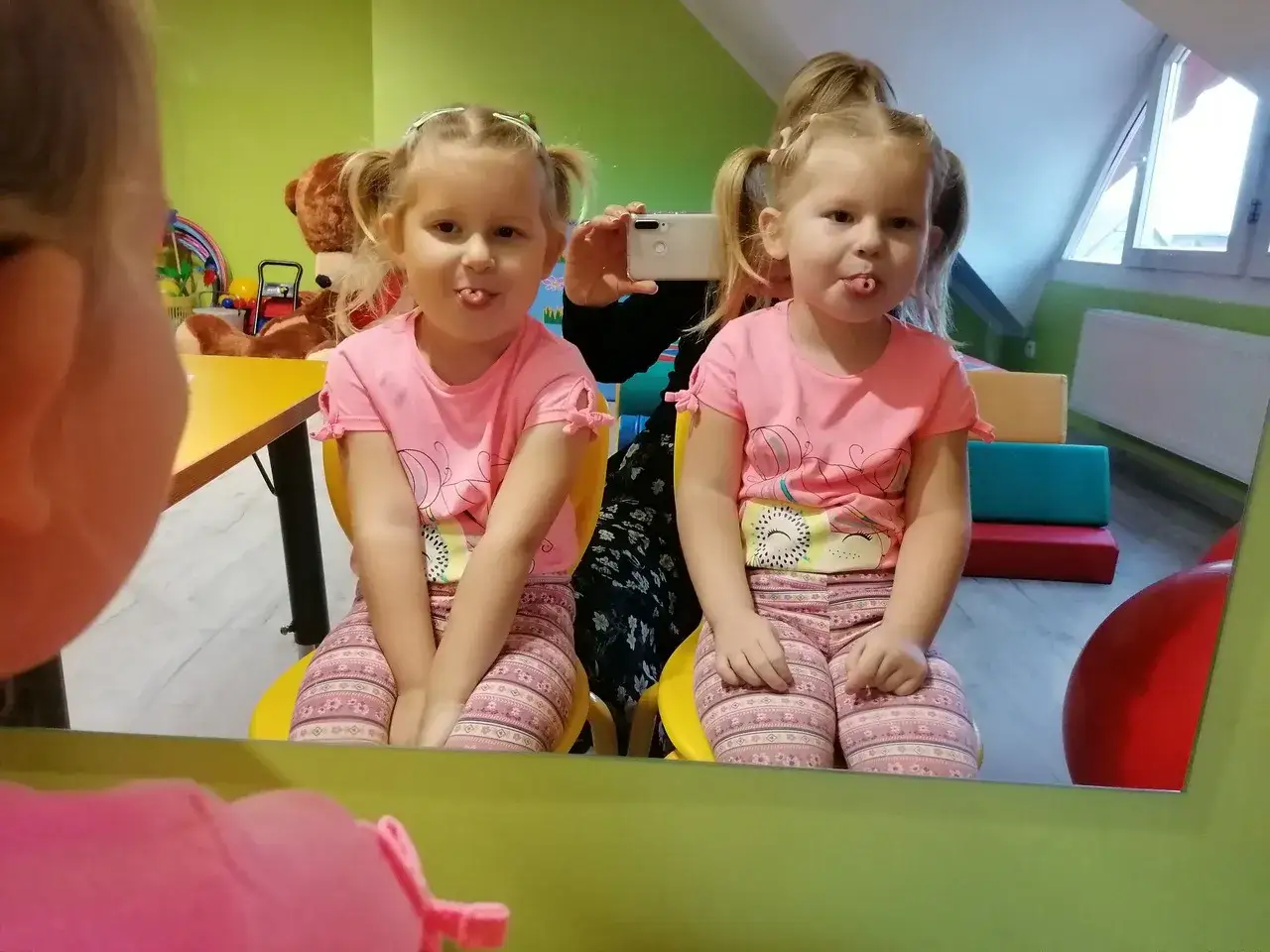 Dwie dziewczynki w różowych bluzeczkach ćwiczą język, wyciągając go do przodu. Wesołe ćwiczenia logopedyczne w sali zabaw.